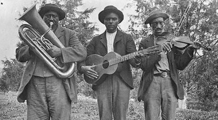 Three African American men are standing holding instruments - a tuba, guitar, and fiddle with a bow. They are each wearing a shirt, pants, coat, and a hat. They are standing outside with trees in the background. Photo: Blount County Public Library
