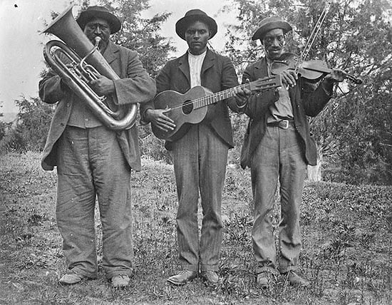 Three African American men are standing holding instruments - a tuba, guitar, and fiddle with a bow. They are each wearing a shirt, pants, coat, and a hat. They are standing outside with trees in the background. Photo: Blount County Public Library